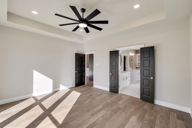 unfurnished bedroom featuring ensuite bath, a raised ceiling, light wood-type flooring, and ceiling fan