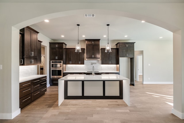 kitchen with an island with sink, light wood-type flooring, hanging light fixtures, stainless steel double oven, and decorative backsplash