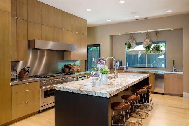 kitchen with a center island with sink, designer stove, light wood-type flooring, range hood, and a breakfast bar