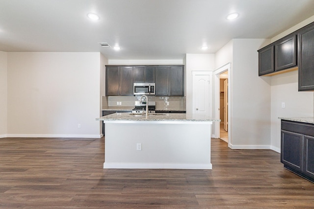 kitchen featuring dark wood-type flooring, appliances with stainless steel finishes, light stone counters, and an island with sink