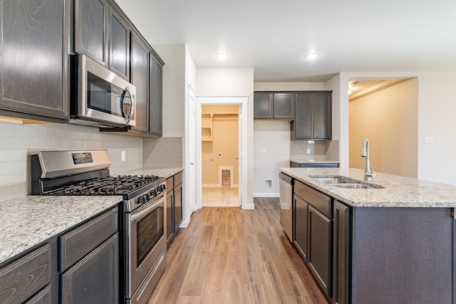 kitchen featuring appliances with stainless steel finishes, an island with sink, sink, light wood-type flooring, and light stone counters