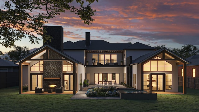 back house at dusk featuring a lawn, an outdoor stone fireplace, a balcony, and a patio