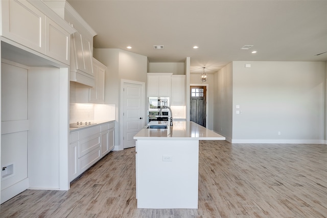 kitchen with black electric stovetop, white cabinets, light hardwood / wood-style floors, stainless steel microwave, and an island with sink