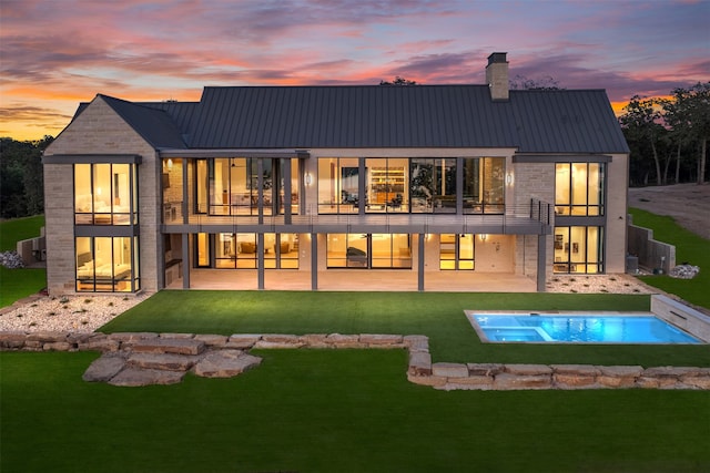 back house at dusk featuring an in ground hot tub, a balcony, a patio area, and a lawn