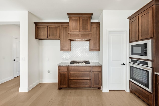 kitchen with backsplash, premium range hood, light wood-type flooring, light stone counters, and stainless steel appliances