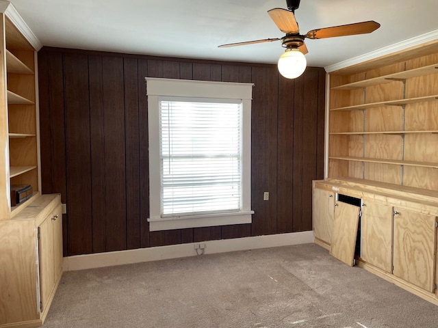 unfurnished living room featuring wood walls, crown molding, ceiling fan, built in features, and light colored carpet