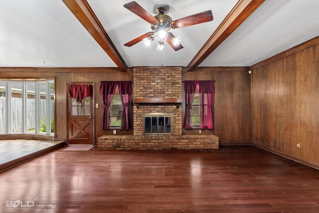 unfurnished living room with a brick fireplace, wooden walls, beam ceiling, dark wood-type flooring, and ceiling fan