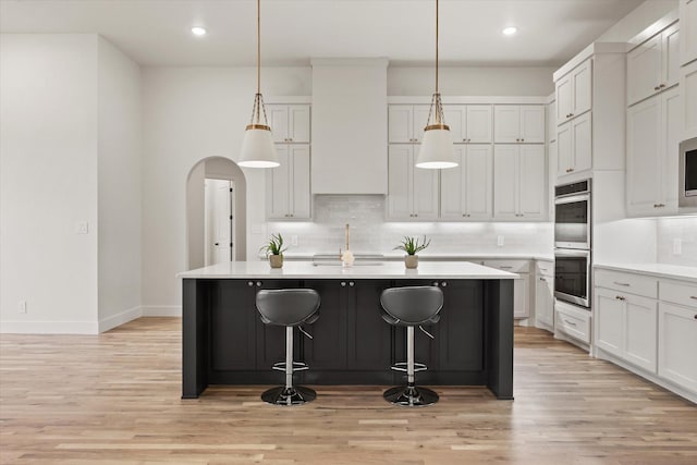 kitchen featuring a kitchen island with sink, white cabinets, double oven, and decorative light fixtures