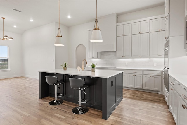 kitchen with pendant lighting, sink, light hardwood / wood-style flooring, a kitchen island with sink, and backsplash