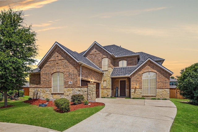 french country inspired facade with stone siding, a yard, an attached garage, a shingled roof, and brick siding