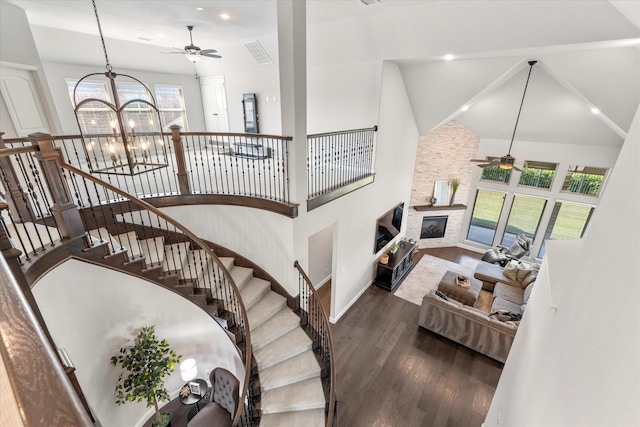 stairs featuring hardwood / wood-style flooring, a stone fireplace, high vaulted ceiling, and ceiling fan with notable chandelier