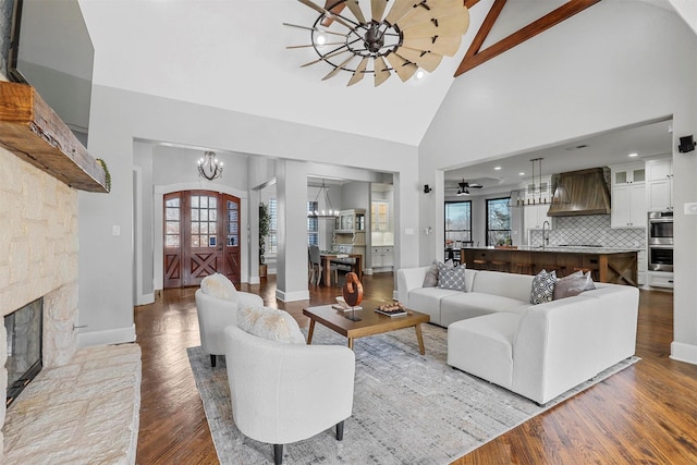 living room featuring a stone fireplace, sink, high vaulted ceiling, light wood-type flooring, and ceiling fan with notable chandelier