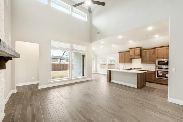 kitchen featuring a stone fireplace, an island with sink, a high ceiling, ceiling fan, and stainless steel appliances