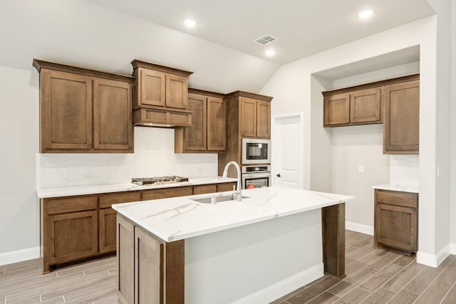 kitchen featuring sink, a kitchen island with sink, stainless steel appliances, light stone countertops, and vaulted ceiling