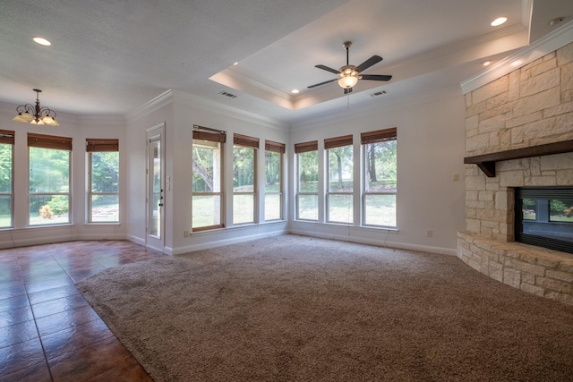 unfurnished living room featuring a raised ceiling, a stone fireplace, dark colored carpet, ceiling fan with notable chandelier, and ornamental molding