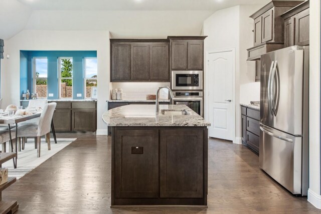 kitchen with appliances with stainless steel finishes, lofted ceiling, a kitchen island with sink, light stone countertops, and dark brown cabinets