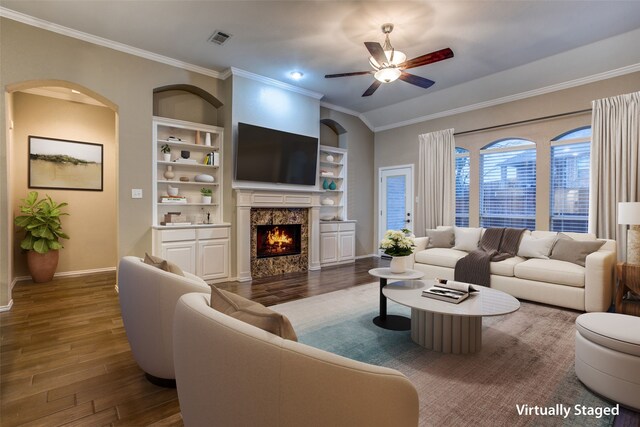 living room featuring a fireplace, ceiling fan, built in shelves, ornamental molding, and dark wood-type flooring