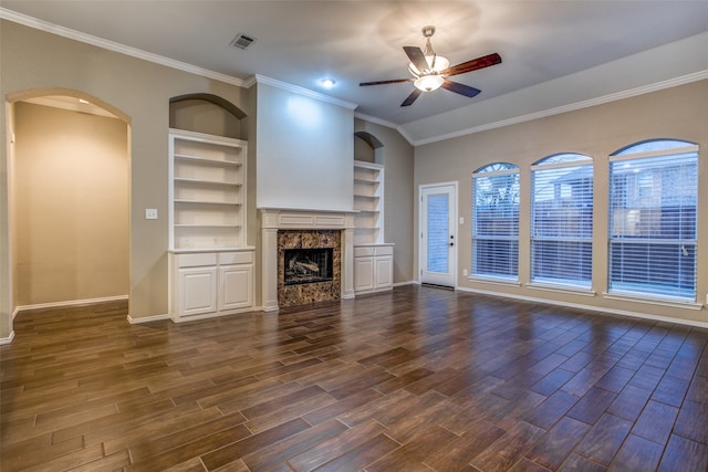 unfurnished living room featuring built in features, a premium fireplace, dark wood-type flooring, ceiling fan, and ornamental molding