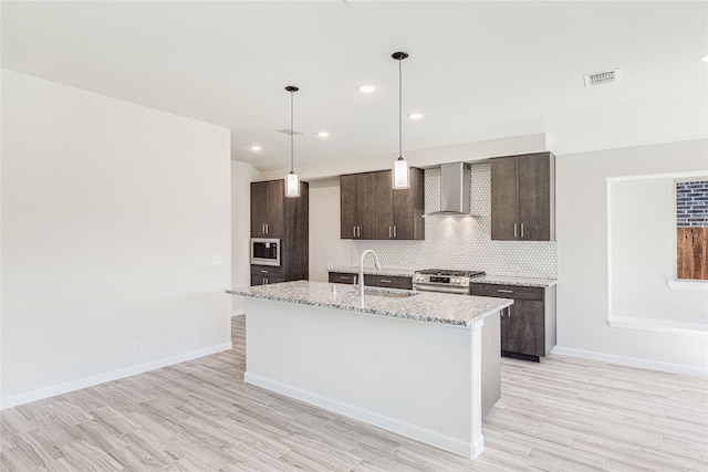 kitchen featuring a kitchen island with sink, backsplash, stainless steel appliances, dark brown cabinetry, and wall chimney exhaust hood