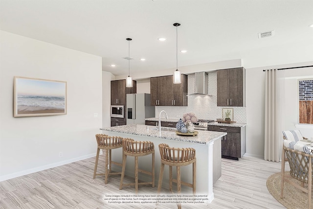 kitchen featuring tasteful backsplash, dark brown cabinets, an island with sink, stainless steel appliances, and wall chimney range hood