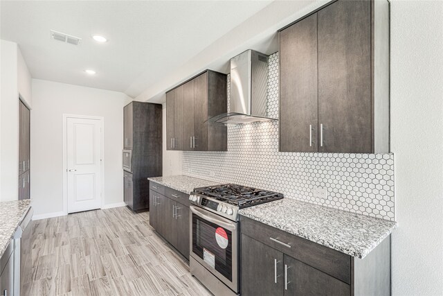 kitchen with backsplash, light stone counters, dark brown cabinetry, stainless steel gas range oven, and wall chimney exhaust hood