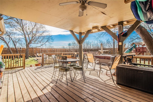 wooden terrace featuring a ceiling fan, outdoor dining space, a fenced backyard, and a playground