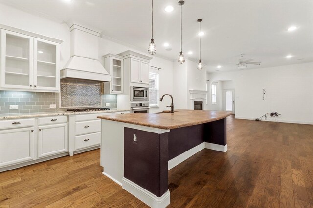 kitchen with premium range hood, decorative backsplash, an island with sink, ceiling fan, and wood-type flooring