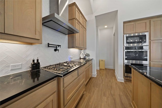 kitchen featuring dark stone counters, stainless steel appliances, wall chimney range hood, light hardwood / wood-style floors, and decorative backsplash