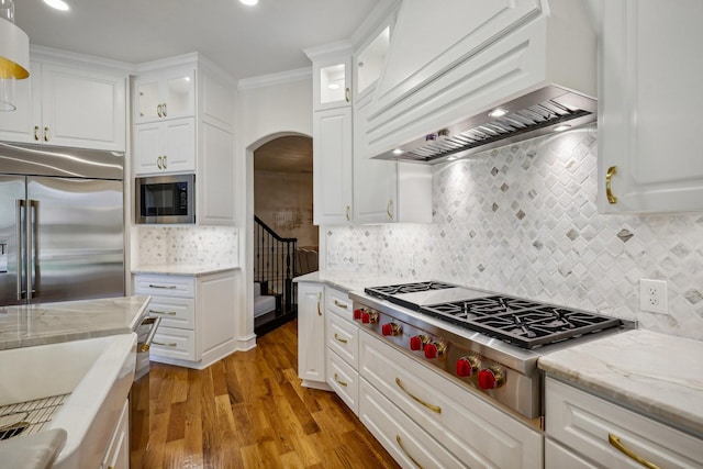 kitchen with white cabinetry, ornamental molding, built in appliances, custom range hood, and light hardwood / wood-style flooring