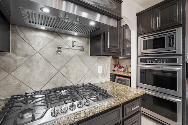 kitchen with decorative backsplash, dark brown cabinets, stainless steel appliances, light stone countertops, and custom range hood