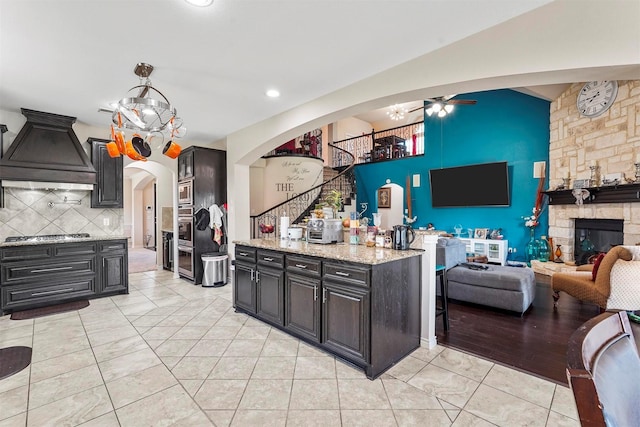 kitchen with premium range hood, a stone fireplace, tasteful backsplash, light stone counters, and ceiling fan