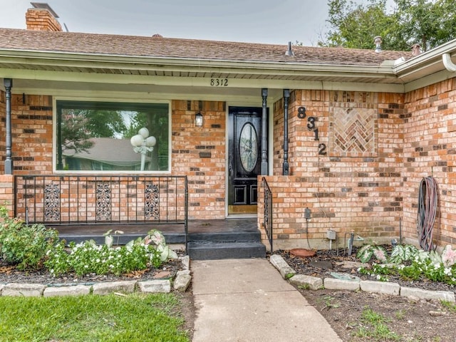 doorway to property featuring covered porch, roof with shingles, brick siding, and a chimney