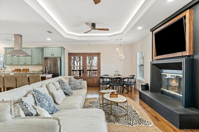 living room featuring french doors, light hardwood / wood-style flooring, ceiling fan, a raised ceiling, and ornamental molding