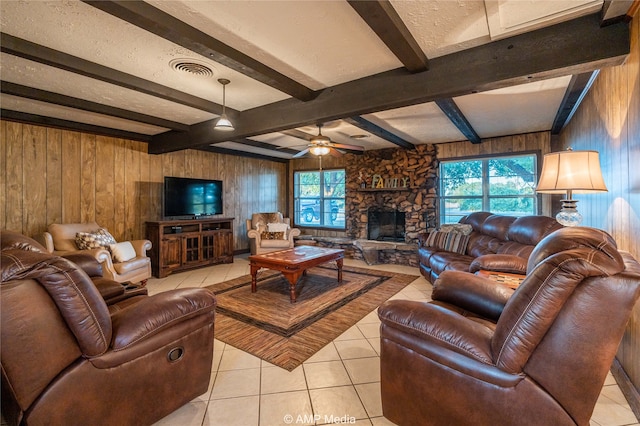 living room featuring light tile patterned flooring, ceiling fan, beam ceiling, wood walls, and a stone fireplace