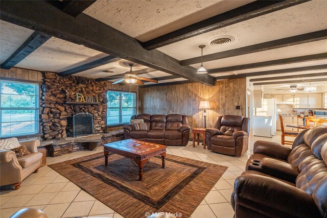 tiled living room with a stone fireplace, wooden walls, ceiling fan, and beam ceiling