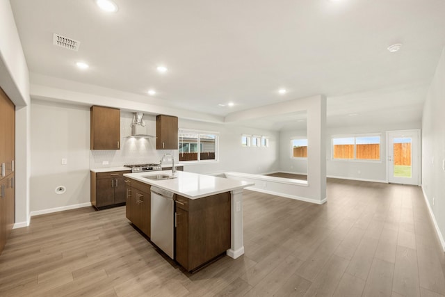 kitchen featuring dishwasher, a kitchen island with sink, sink, wall chimney exhaust hood, and light hardwood / wood-style floors