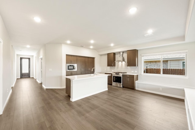 kitchen with appliances with stainless steel finishes, wall chimney exhaust hood, sink, wood-type flooring, and a center island with sink