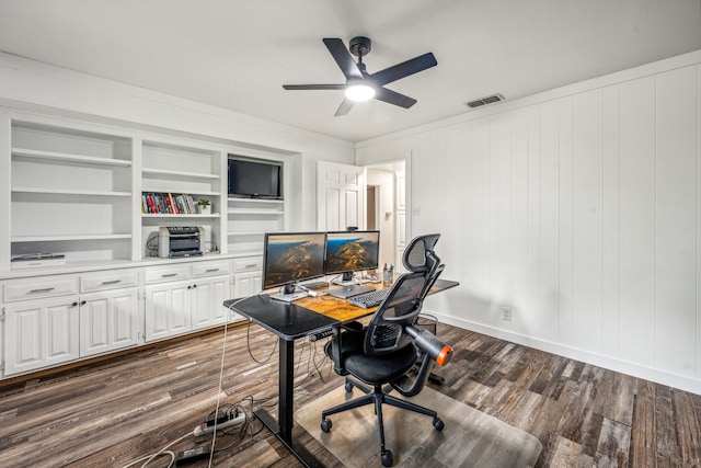 office space featuring built in shelves, ceiling fan, dark wood-type flooring, wooden walls, and ornamental molding