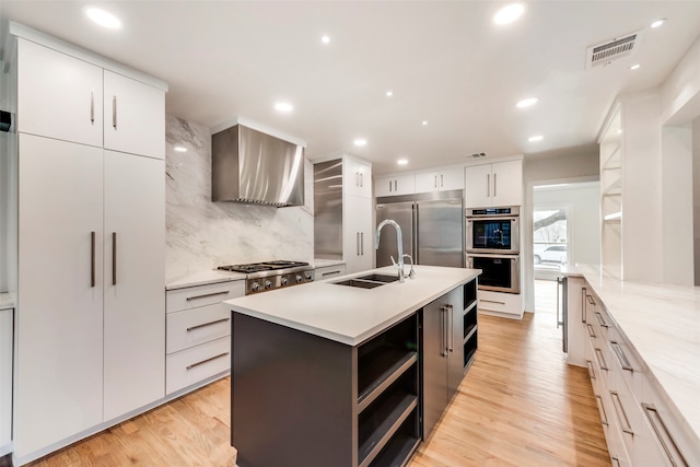 kitchen featuring a kitchen island with sink, stainless steel appliances, sink, white cabinets, and wall chimney range hood