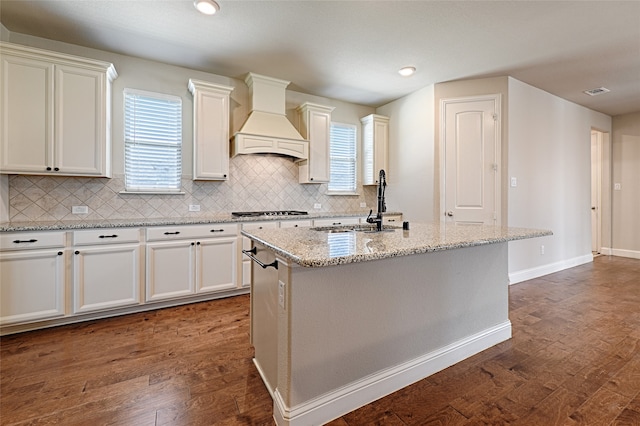 kitchen featuring custom exhaust hood, tasteful backsplash, dark hardwood / wood-style flooring, light stone countertops, and a kitchen island with sink