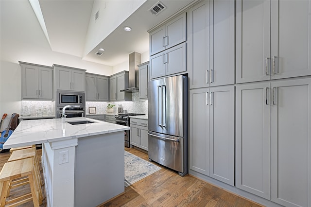 kitchen featuring gray cabinets, a kitchen bar, stainless steel appliances, light stone countertops, and wall chimney range hood