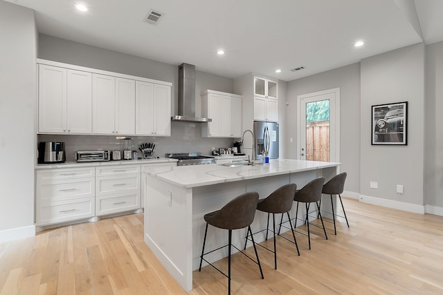 kitchen featuring wall chimney exhaust hood, an island with sink, sink, white cabinetry, and appliances with stainless steel finishes