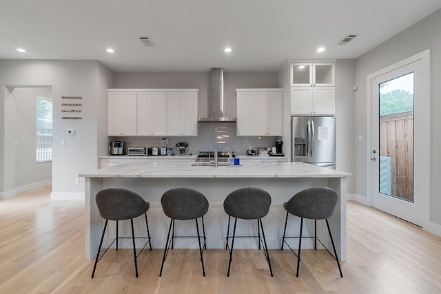 kitchen with white cabinetry, a large island with sink, wall chimney range hood, and stainless steel fridge