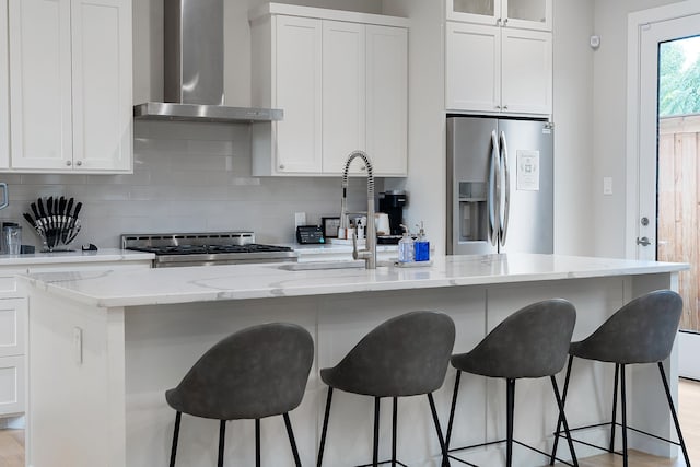 kitchen with wall chimney exhaust hood, appliances with stainless steel finishes, a kitchen island with sink, and white cabinetry