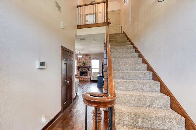 stairs featuring ceiling fan, brick wall, dark hardwood / wood-style floors, a brick fireplace, and a towering ceiling