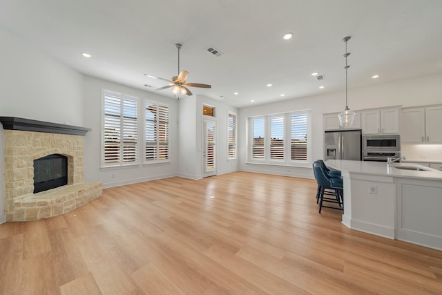 unfurnished living room with ceiling fan, sink, a stone fireplace, and light hardwood / wood-style floors