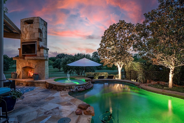 pool at dusk with an in ground hot tub, an outdoor stone fireplace, and a patio area