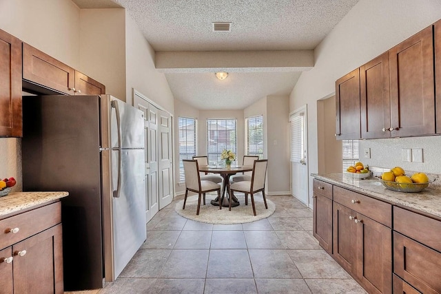 kitchen featuring vaulted ceiling with beams, light tile patterned floors, light stone countertops, a textured ceiling, and stainless steel refrigerator