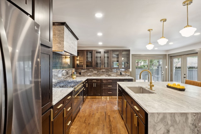 kitchen featuring a kitchen island with sink, custom range hood, hanging light fixtures, sink, and appliances with stainless steel finishes