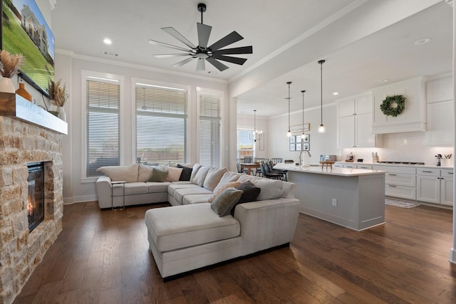 living room featuring a stone fireplace, dark hardwood / wood-style floors, sink, ceiling fan, and crown molding